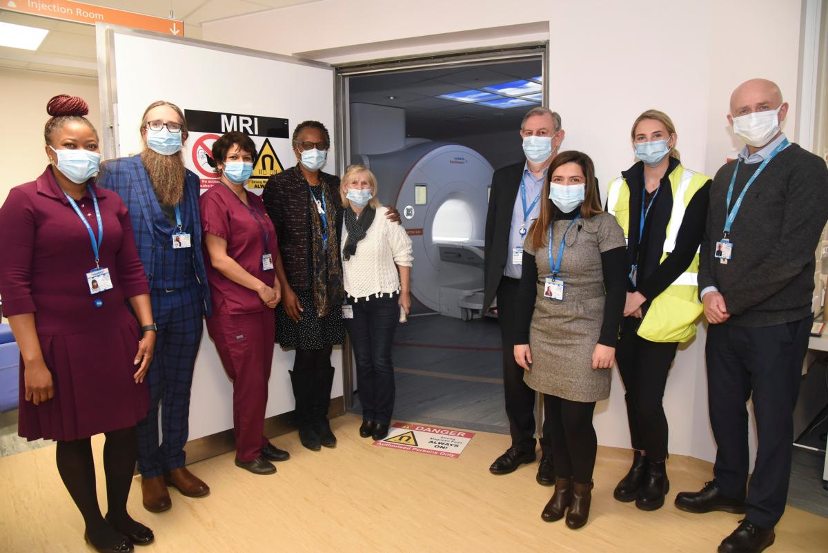 approximately eight members of NHS staff, some in burgundy uniform tunics, standing in front of a doorway labeled 'MRI' through which a large body scanning machine can be seen
